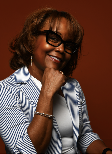 Karen smiling with her head tilted slightly to her left and her chin propped up by her right hand. She's wearing glasses with black frames with blue and white striped blazer and white tee underneath.