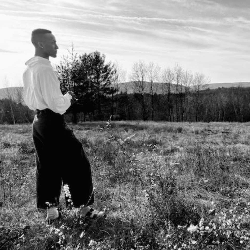 Young black man, in a field, with a loose fitting white button down shirt and baggy black slacks looking forward at the landscape.