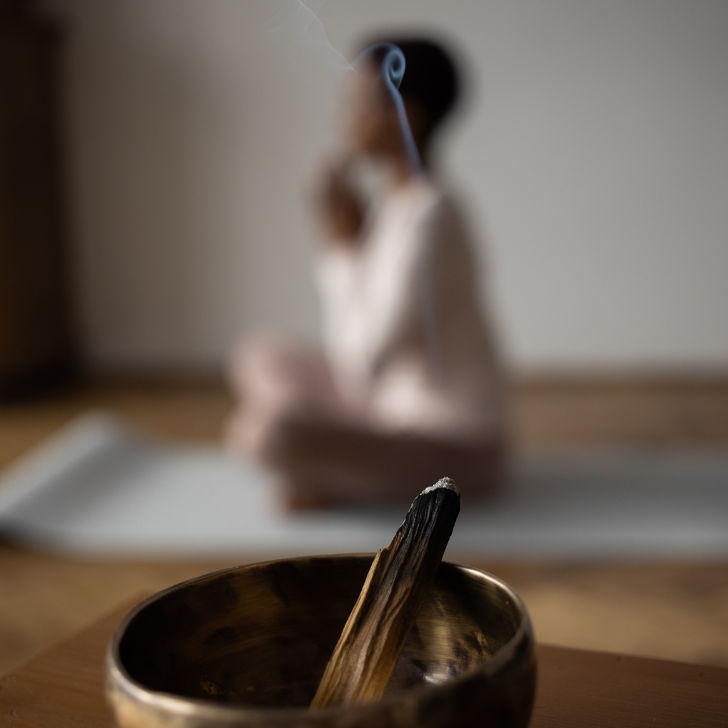 In the background, a black women in a mediation pose. In the foreground, a pewter bow burning incense or sage.